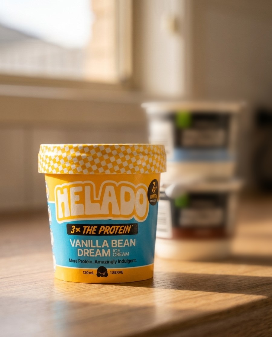 A sealed Helado Vanilla Bean Dream cup sharp in the foreground on a sunlit kitchen bench, two unbranded supermarket protein dessert pots out of focus behind it