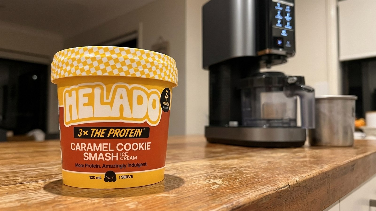 A sealed Helado Caramel Cookie Smash cup on a kitchen bench, a countertop ice cream maker and its washed canister behind it