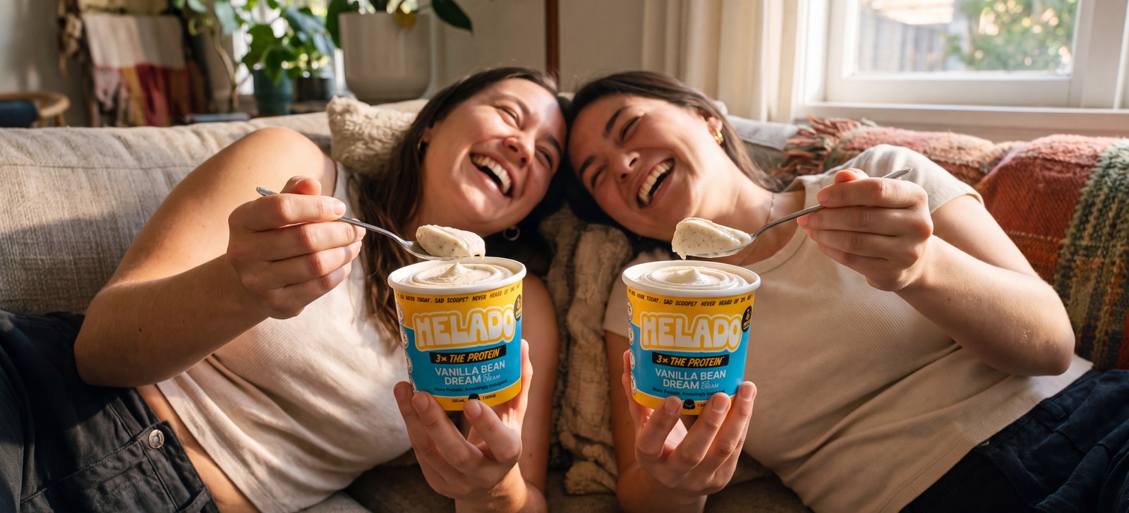 Two friends laughing together on a couch, each holding an open Helado cup with a soft vanilla swirl above the rim